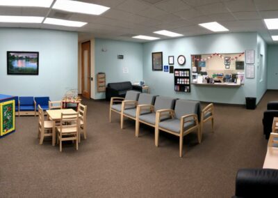 picture of office lobby and waiting area with chairs, side tables, toys and play area in one corner, light blue walls, reception window, clock and artwork on walls