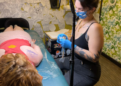 Individual laying on their stomach to receive a tattoo on their back. Tattoo artist sitting on a stool wearing gloves and a face mask and holding a tattoo machine at Glory Bound Tattoo.