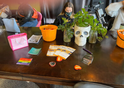 MOCSA stickers and brochures sitting on a table with plants and other items. Individuals wearing facemasks sitting behind table not looking at camera.