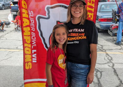 An adult and child at MOCSA's Chiefs tailgate party standing in front of a Chiefs flag smiling at the camera.