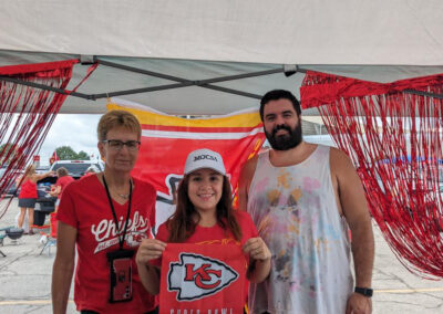 Three individuals at MOCSA's Chiefs tailgate party standing in front of Chiefs flag and smiling at the camera.