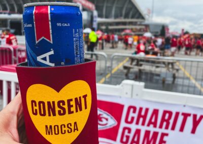 close up image of a hand holding a red can coozie with gold heart and the word CONSENT. Blurry background of Arrowhead stadium and Chiefs Charity Game banner.